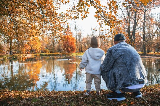Grandfather and small child standing by a calm lake in an autumn park. Concept of authenticity, family time, love and connection with nature..