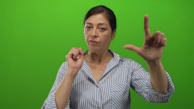Middle aged hispanic woman making loser hand sign with raised thumb and index finger in green studio setting; mockery taunt.