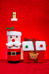 a wine bottle dressed in a festive Santa Claus-themed knitted cover and a present box on red at vertical composition