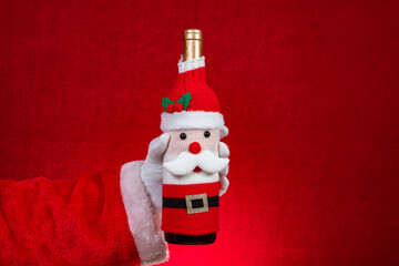 Santa Claus hand holding a wine bottle dressed in a festive Santa Claus-themed knitted cover on red at horizontal composition