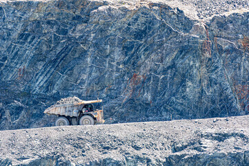 Large mining dump truck loaded with rocks drives along a road inside an open pit quarry. Industrial heavy machinery working in a rugged geological environment under daylight.
