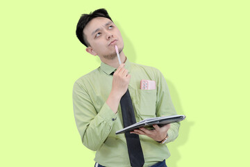 A pensive young Asian professional holding a pen with a deep, concentrated expression while holding a notepad, posing or deep thought against a light green background