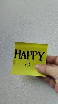 Una toma de cerca de una mano de mujer con una nota adhesiva de color amarillo con la palabra feliz  en movimiento en un fondo blanco. Video vertical
