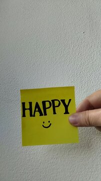 Una toma de cerca de una mano de mujer con una nota adhesiva de color amarillo con la palabra feliz  en movimiento en un fondo blanco. Video vertical