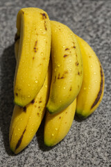  Close-up photo of ripe yellow bananas with natural brown spots and fresh water droplets resting on a gray stone surface.
