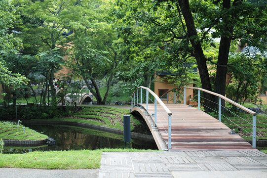 A modern wooden footbridge with sleek metal railings arches gently over a narrow water channel. The scene is immersed in a lush green environment with tall, leafy trees and manicured grassy banks