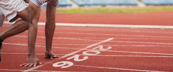 A runner crouches at the starting line of a track, getting ready to sprint. The clean red surface and numbered lanes reveal an active sports environment on a bright day.