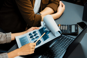 Cropped working asian smart manager with coworker discussing paperwork comparing business data report on laptop at modern home office, taking note with customer segment to marketing plan. Infobahn.