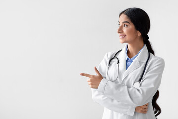A smiling Indian woman doctor stands in a white coat, pointing to an empty space. She is a family therapist ready to share important healthcare information in a studio setting.