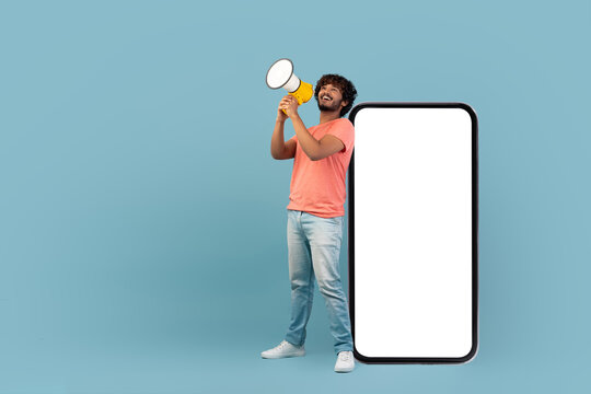A positive young Indian man with curly hair holds a megaphone while standing next to a large smartphone featuring a blank screen. He smiles as he makes an announcement in a blue studio.