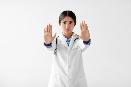 Serious young Indian lady doctor in a white coat holds up her hands in a stop gesture against a white background. She emphasizes the importance of safety and social distancing during COVID-19.