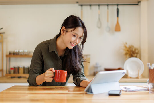 Asian woman drink a cup of coffee and reading news on digital tablet in a kitchen using laptop at home, Activity lifestyles concept
