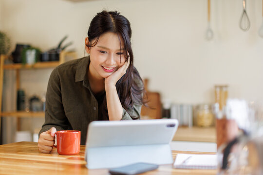 Asian woman drink a cup of coffee and reading news on digital tablet in a kitchen using laptop at home, Activity lifestyles concept