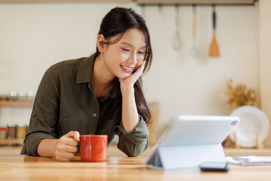 Asian woman drink a cup of coffee and reading news on digital tablet in a kitchen using laptop at home, Activity lifestyles concept
