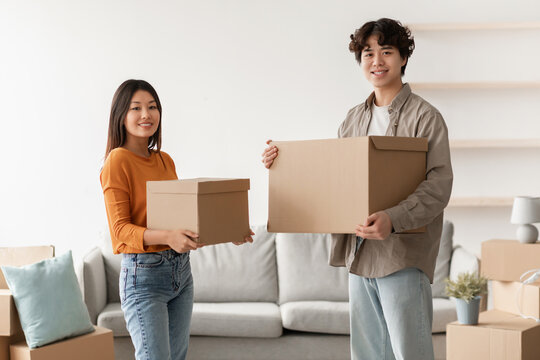 Cheerful young Asian couple stands in their new apartment holding cardboard boxes. They are smiling as they enjoy their moving day and the excitement of starting a new chapter together.