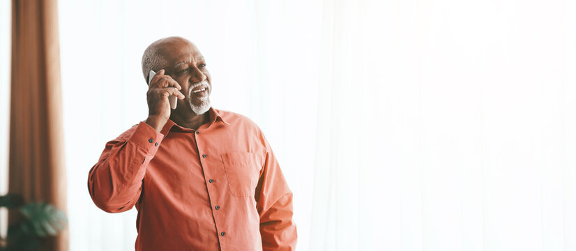 A senior man wearing a bright orange shirt smiles as he communicates on his phone, standing by a large window that lets in plenty of light, creating a cheerful atmosphere.