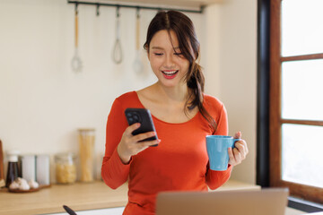 Happy asian woman drinking tea while using smart phone in the kitchen. Copy space.