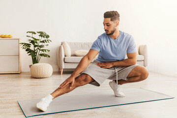 A man in a light blue shirt and shorts is performing a stretch on a yoga mat in a sunlit living room. A couch and a plant are visible in the background, creating a relaxing atmosphere.