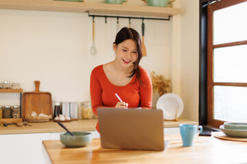 A asian eastern young woman happily jotting down notes while looking at her laptop at a kitchen counter, studying online
