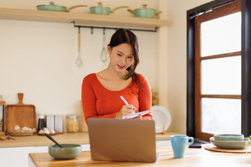 A asian eastern young woman happily jotting down notes while looking at her laptop at a kitchen counter, studying online
