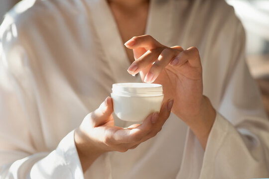 Close up of female hands applying beauty care product while wearing a silk bathrobe. The woman engages in her morning routine after a refreshing shower, holding a jar of cream.