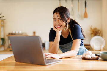 Interested woman look into laptop screen, choosing film, program, movie, video to watch while cooking in kitchen. asian female reading food recipes in internet, surfing in social networks, web pages
