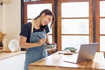 Woman, cooking in kitchen with laptop for live streaming, online recipe and meal prep by stove. Nutrition influencer, food blogger or vegan chef with social media tutorial in apartment
