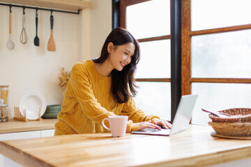 Smiling asian female freelancer is working from home, sitting at a table in her modern apartment, using a laptop 
