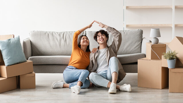 Two joyful millennial Asian individuals sit on the floor among carton boxes in their new home, making a house roof gesture. It is a special moment on their moving day filled with excitement. - Powered by Adobe