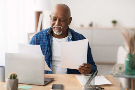 A senior African businessman is seated at his desk, attentively reviewing paperwork while using a laptop in a bright, contemporary office environment. He appears focused on his tasks.