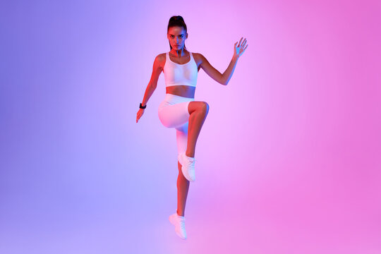 A fitness enthusiast executes a standing crunch by raising her knee in a neon pink and blue studio. She looks confidently at the camera while working on her cardio routine. - Powered by Adobe
