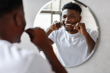 A cheerful black man stands in a modern bathroom, flossing his teeth while looking in the mirror. He enjoys his daily oral hygiene routine in bright morning light. © Prostock-studio