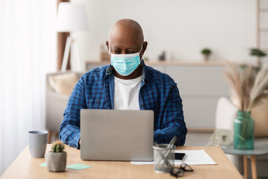 A mature Black man wearing a protective face mask is focused on his laptop while sitting at a desk in a modern office. This scene reflects remote work during the Covid-19 pandemic. - Powered by Adobe