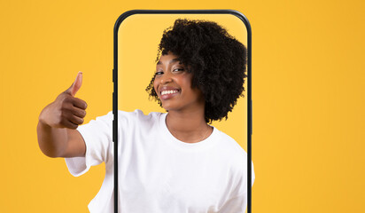 A young woman smiles and gives a thumbs up while posing in front of a vibrant yellow backdrop. The...