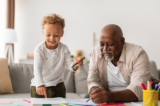 Grandfather and grandson are happily sketching together at a table in their cozy home. They are using colorful markers, sharing laughs and making memories during their weekend activity.