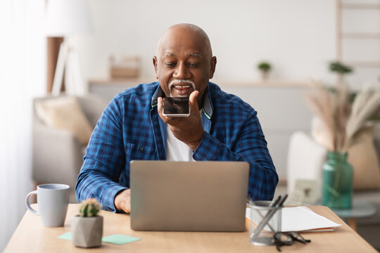 Senior African American man engages with a voice search application on his smartphone while seated at a laptop. He works in a bright, modern office environment surrounded by plants and stationery.