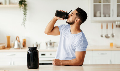 A fit man is drinking a protein shake from a black shaker in a well-lit kitchen. He looks relaxed and revitalized after his workout. The background features a clean countertop and cooking utensils.