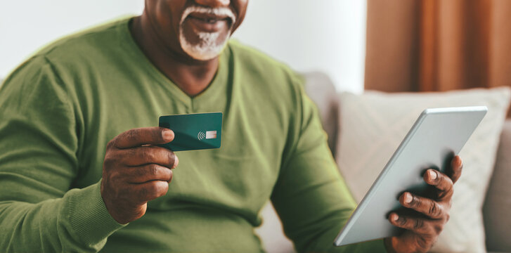 A senior man sits comfortably in a cozy living room, holding a credit card in one hand and a tablet in the other. He appears focused on making an online purchase during daytime.