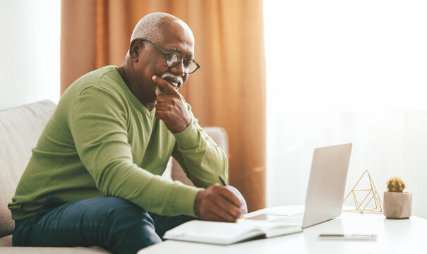 An elderly man studies intently at his home, seated on a couch with a notebook and laptop in front of him. Warm sunlight fills the cozy living room, creating a relaxed atmosphere.