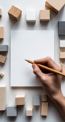 Hand holding pencil over blank notepad surrounded by various wooden cubes