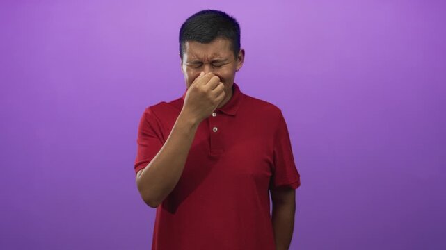 Young hispanic man pinching nose with fingers, eyes closed, wearing red polo shirt in purple studio; disgust reaction.
