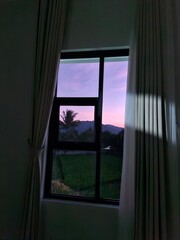 View of a colorful purple and pink sunset sky with a palm tree, seen through a dark room's window.