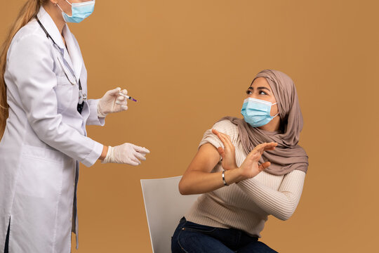 Young arab lady in face mask scared to make coronavirus vaccine injection, making STOP gesture and looking at doctor. Muslim woman opposing immunization against covid-19 - Powered by Adobe