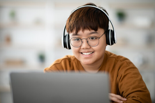 Closeup of a cheerful asian boy with glasses, smiling while using a laptop and wireless headset.