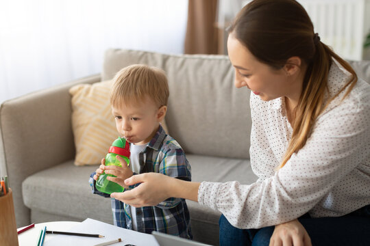 A mother assists her young child with a colorful sippy cup in a cozy living room. They are seated at a table filled with art supplies, creating a warm atmosphere.