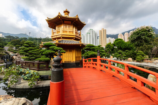 Classic chinese pagoda with arched wooden bridge, in the traditional garden setting of Nan Lian garden, in Hong Kong. Uban skyline in the background; cloudy sky overhead.
 - Powered by Adobe