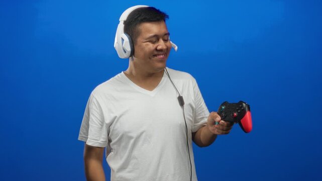 Young hispanic man wearing white t shirt and headset holding a game controller and pointing finger in blue studio; joy gaming fun.