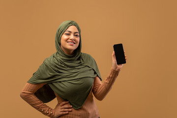 Arab woman stands smiling while holding a smartphone with a blank screen. She wears a green headscarf and a beige top, highlighting her excitement. The background is simple and neutral.