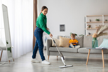A woman in green shirt cleans the living room floor with a mop. Her young child sits on the gray sofa, focused on a tablet, in a cozy, contemporary home environment filled with natural light.