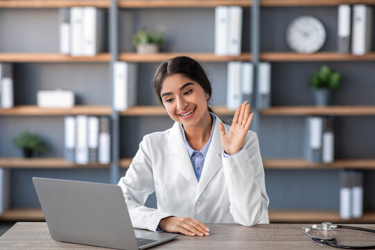 A cheerful young Indian female doctor dressed in a white coat works at her clinic office while waving to her laptop. She engages in a video call meeting with a patient during the covid-19 lockdown.